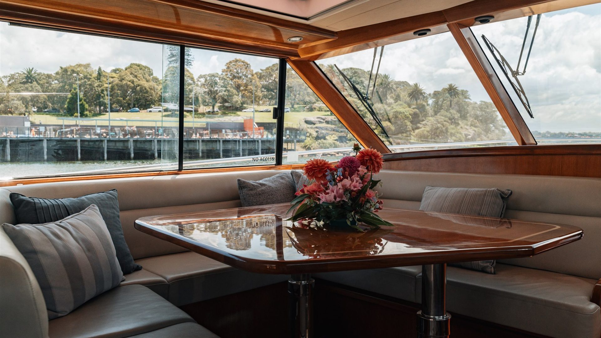 A cozy dining area inside a boat with a polished wooden table decorated with a flower arrangement. The area has cushioned seating with pillows, large windows offering a view of green trees and a waterfront scene with a dock and buildings in the background.