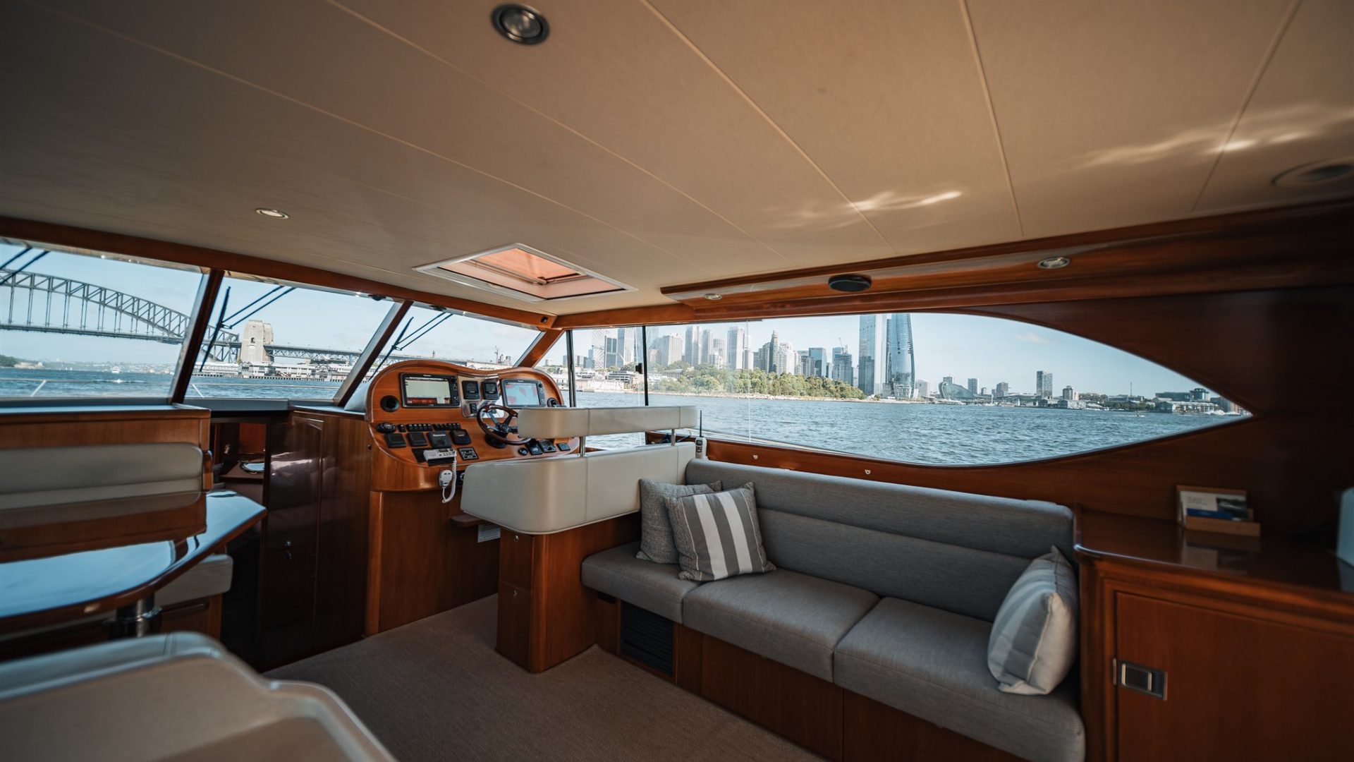 A luxurious boat interior with wooden furnishings and large windows, offering a view of a city skyline with towering buildings and a bridge in the background. There is a comfortable seating area with a grey couch and striped pillows.