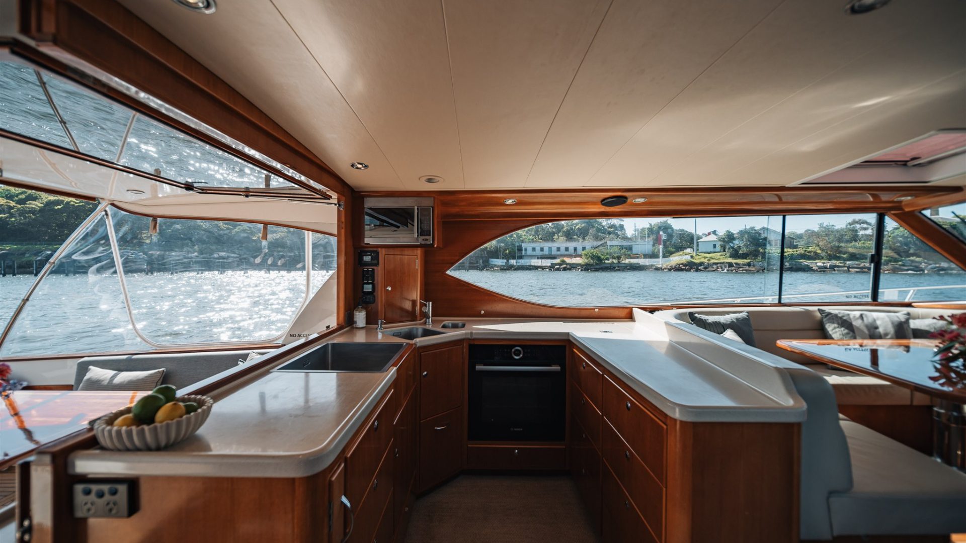 The image shows the interior of a yacht with a modern kitchen. The wooden cabinetry and countertops give it a warm, elegant feel. Large windows offer a view of the water and shoreline with trees and houses. A bowl of fruit and a vase of flowers add a homely touch.