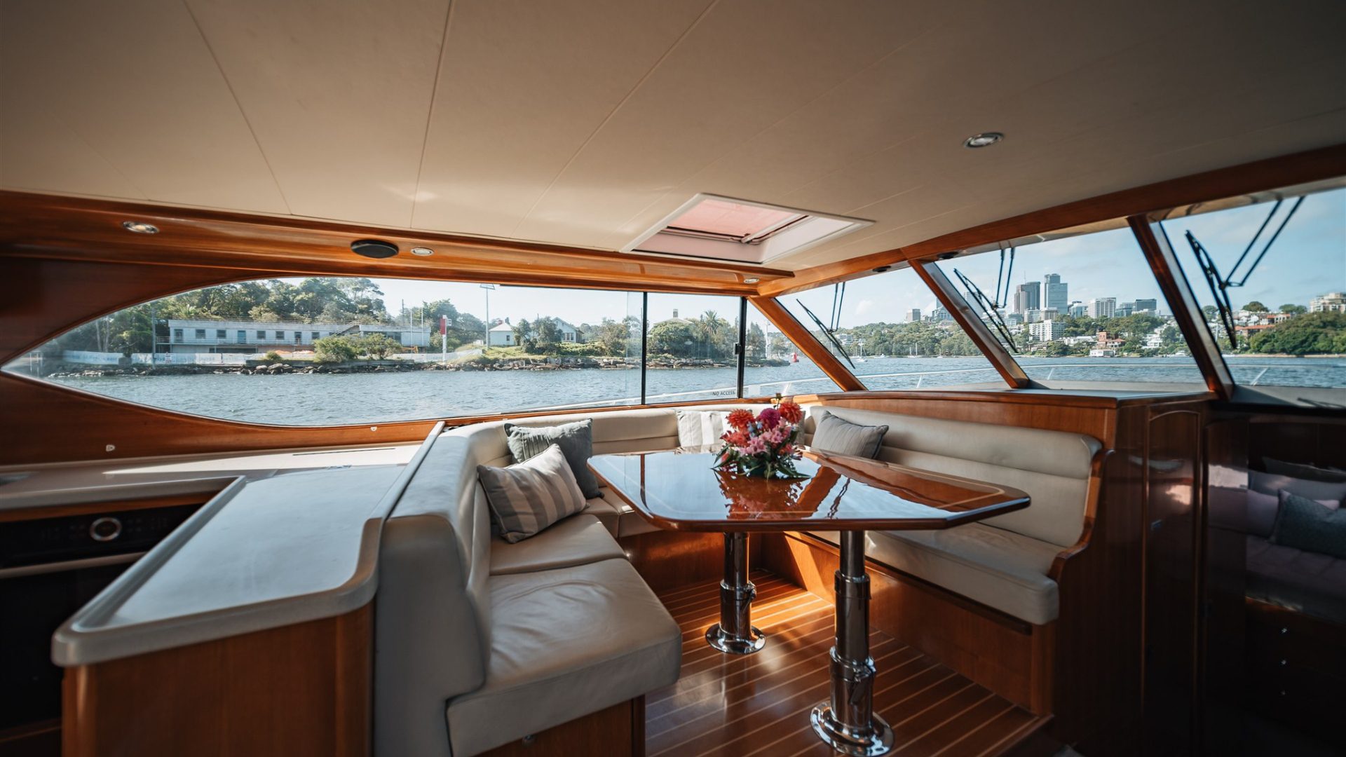Interior of a luxurious boat with a polished wooden table and cushioned seating arranged around it. A bouquet of flowers sits on the table. Large windows offer a view of the water, a nearby green landscape, and a distant city skyline.