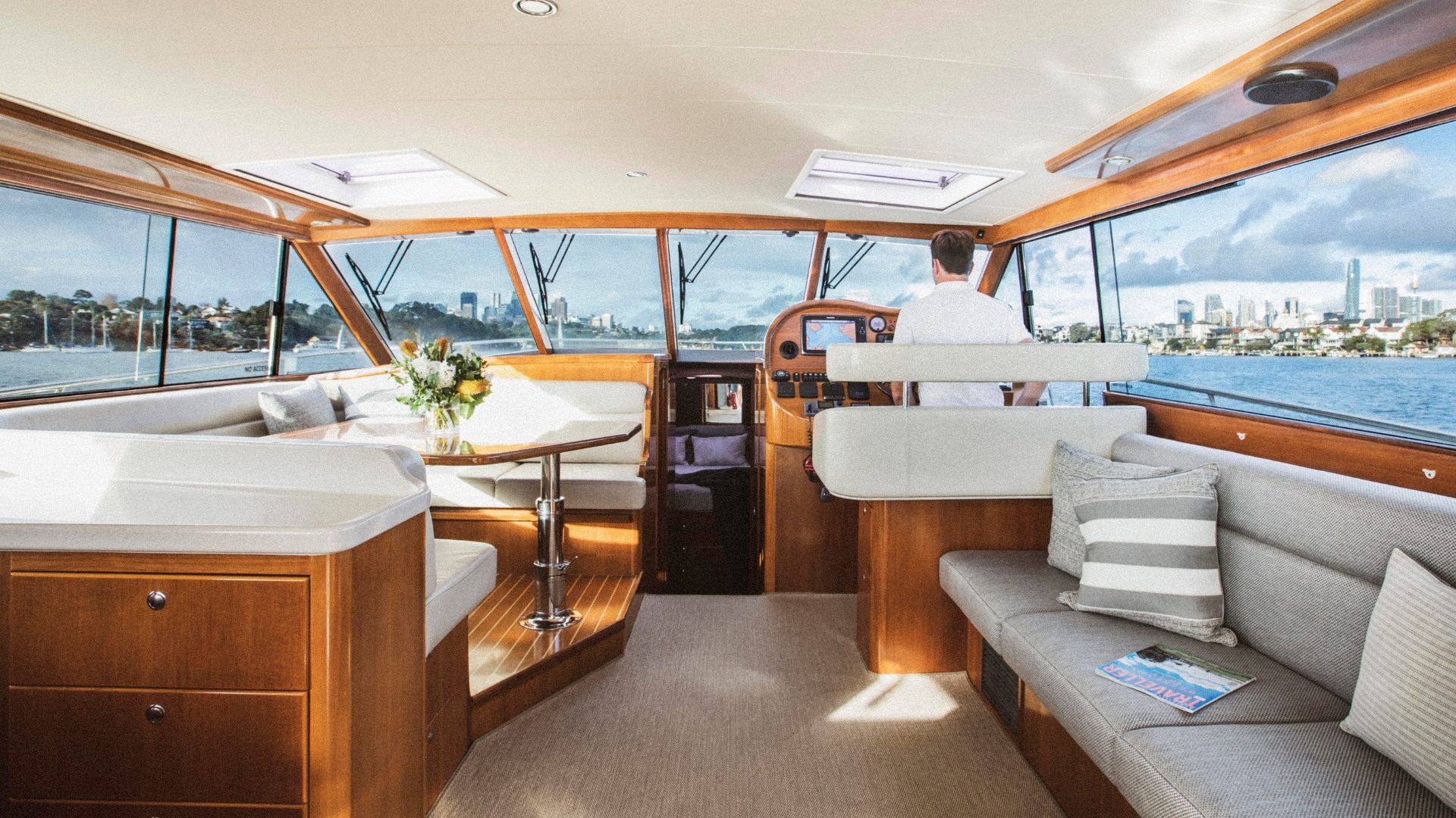 A spacious yacht interior with polished wooden finishes and large windows. It features a white dining area with floral centerpiece on the left, a beige cushioned seating area with striped pillows on the right, and a person standing at the helm steering the boat.