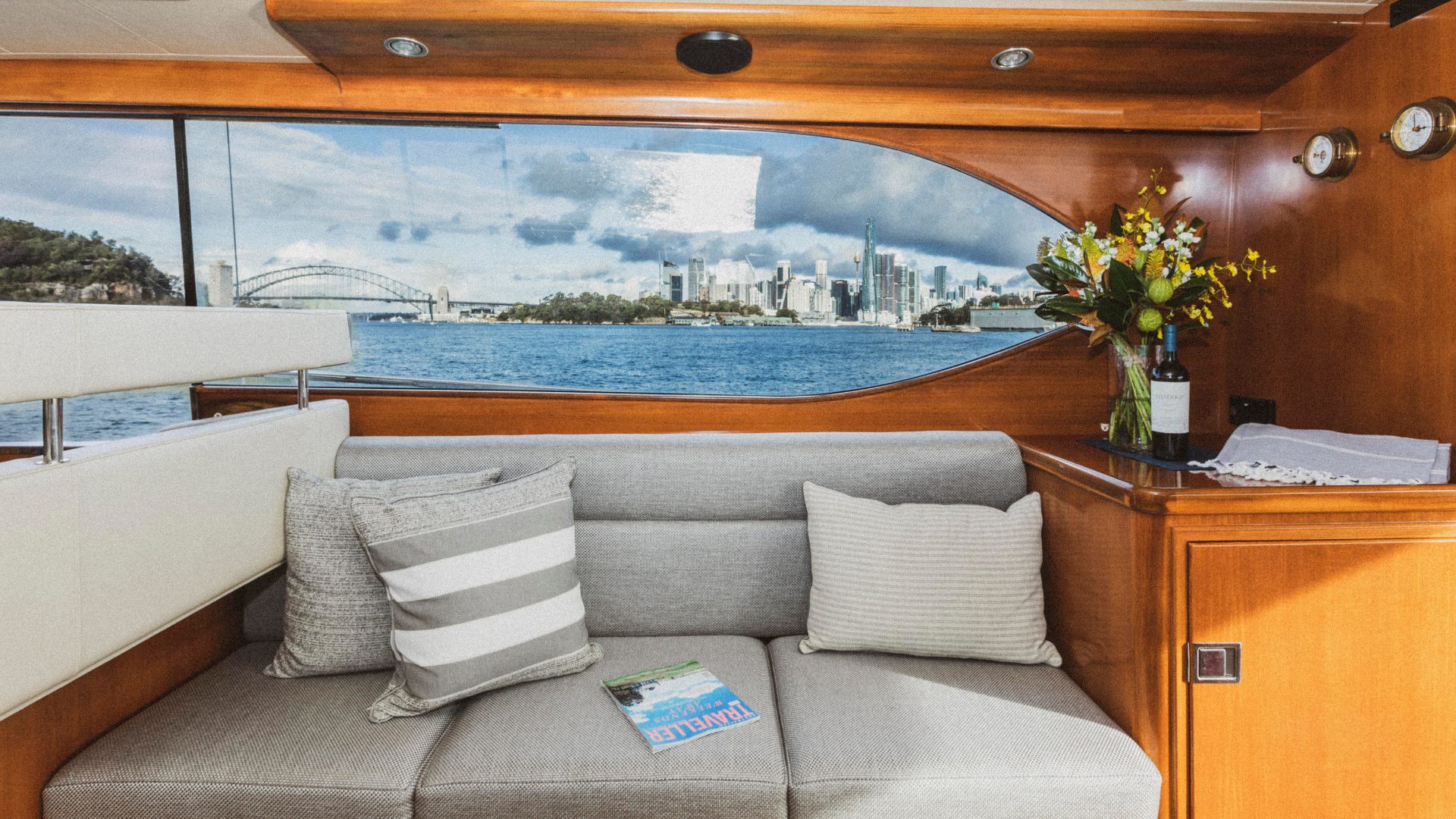 A cozy yacht interior with a gray couch adorned with striped and ribbed cushions. A table on the right holds a vase with a floral arrangement. Large windows reveal a scenic view of a harbor, city skyline, and a bridge under a blue sky with clouds.