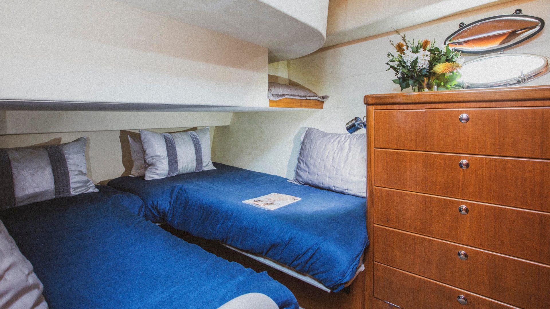 A cozy twin bed cabin with blue bedding and white pillows. A booklet is placed on one bed. There's a wooden dresser with a mirrored knob and a floral arrangement on top. The cabin has a small, oval-shaped window with an open porthole letting daylight in.