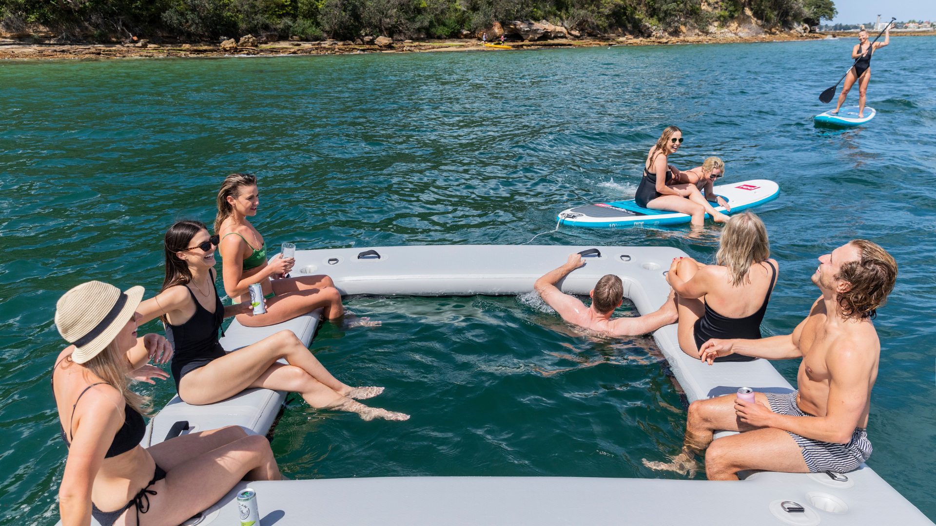 A group of people relaxes on an inflatable square floating structure in the water. Some sit on the edges, chatting and having drinks. Two individuals are on paddleboards nearby. The scene is set on a sunny day with a lush, green shoreline in the background.