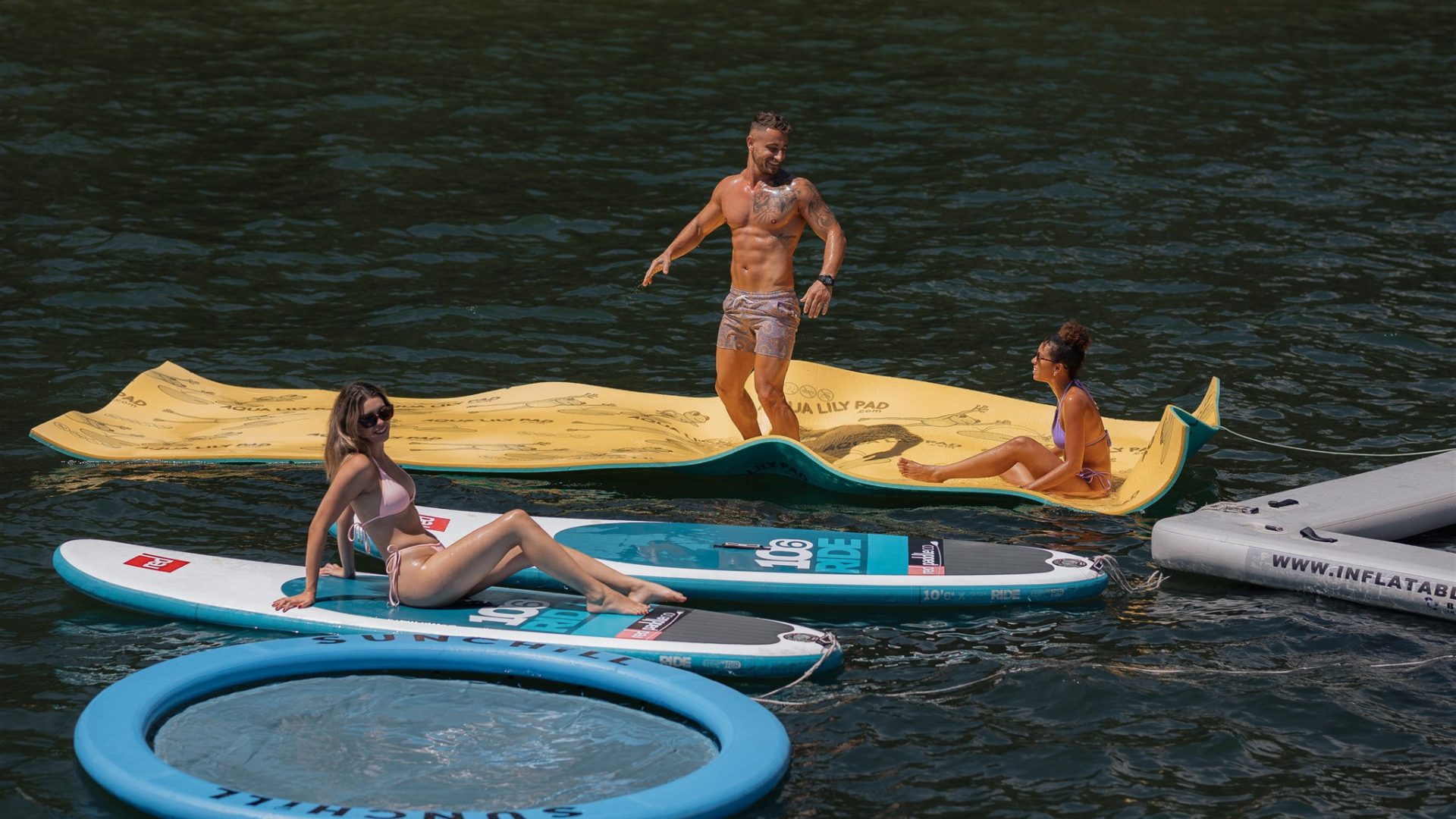 Three people relaxing on water: a woman sitting on a paddleboard, another woman lying on a yellow floating mat, and a shirtless man standing on the mat. They are surrounded by additional floating devices in a calm body of water, enjoying a sunny day.