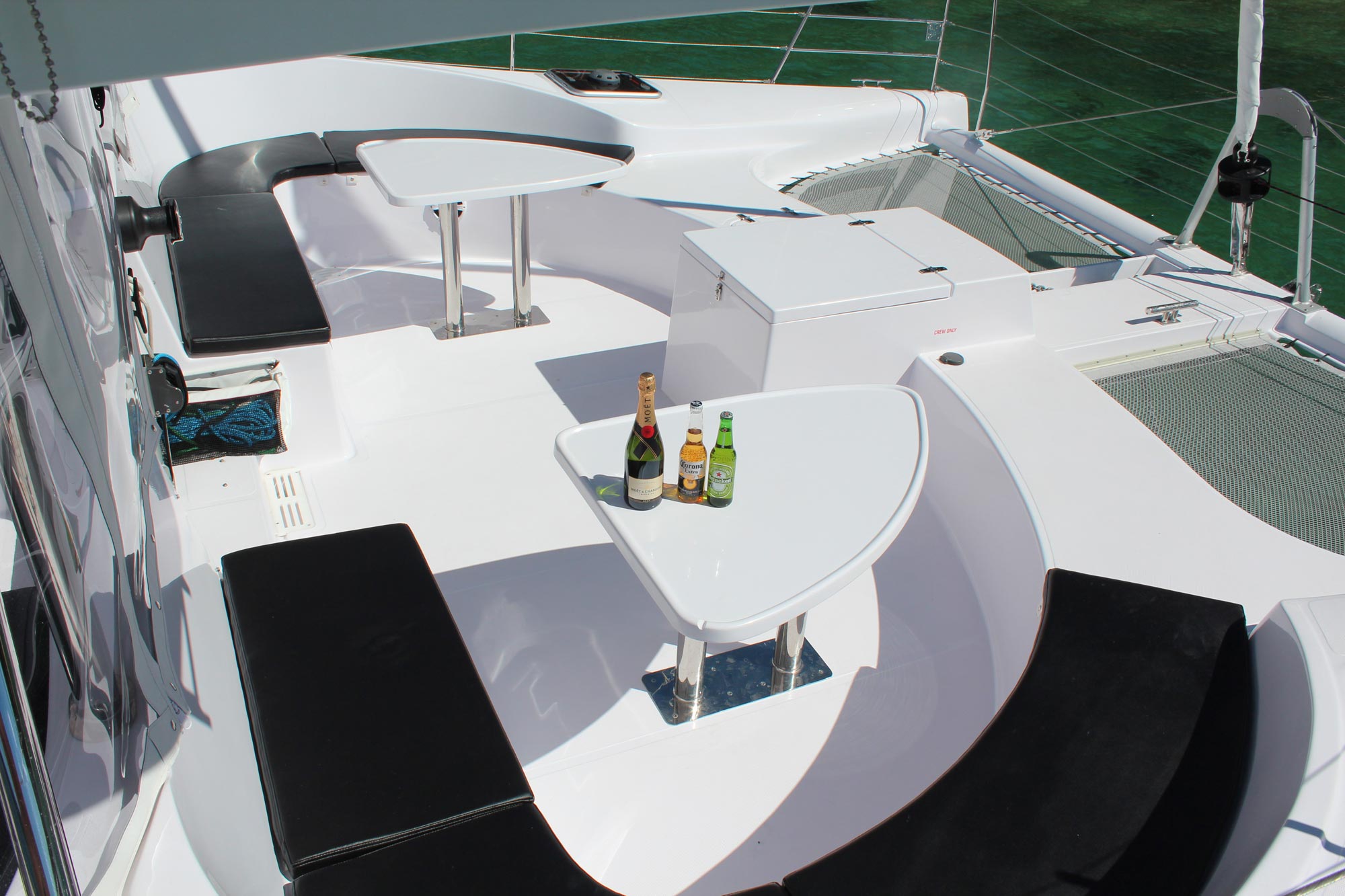 A luxurious yacht deck with white seating and black cushions. The deck has two white tables, each with beverages: one has champagne and the other has two beer bottles. The surrounding waters are clear and green.