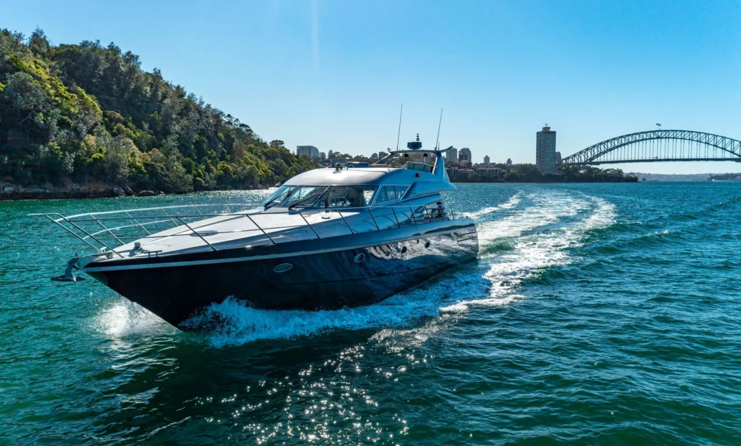A large, sleek motor yacht cruises on clear blue waters near a lush, green coastline. The iconic Sydney Harbour Bridge is visible in the background under a bright, sunny sky. Offered by The Yacht Social Club Sydney Boat Hire, this luxury yacht leaves a trail of white waves as it moves.