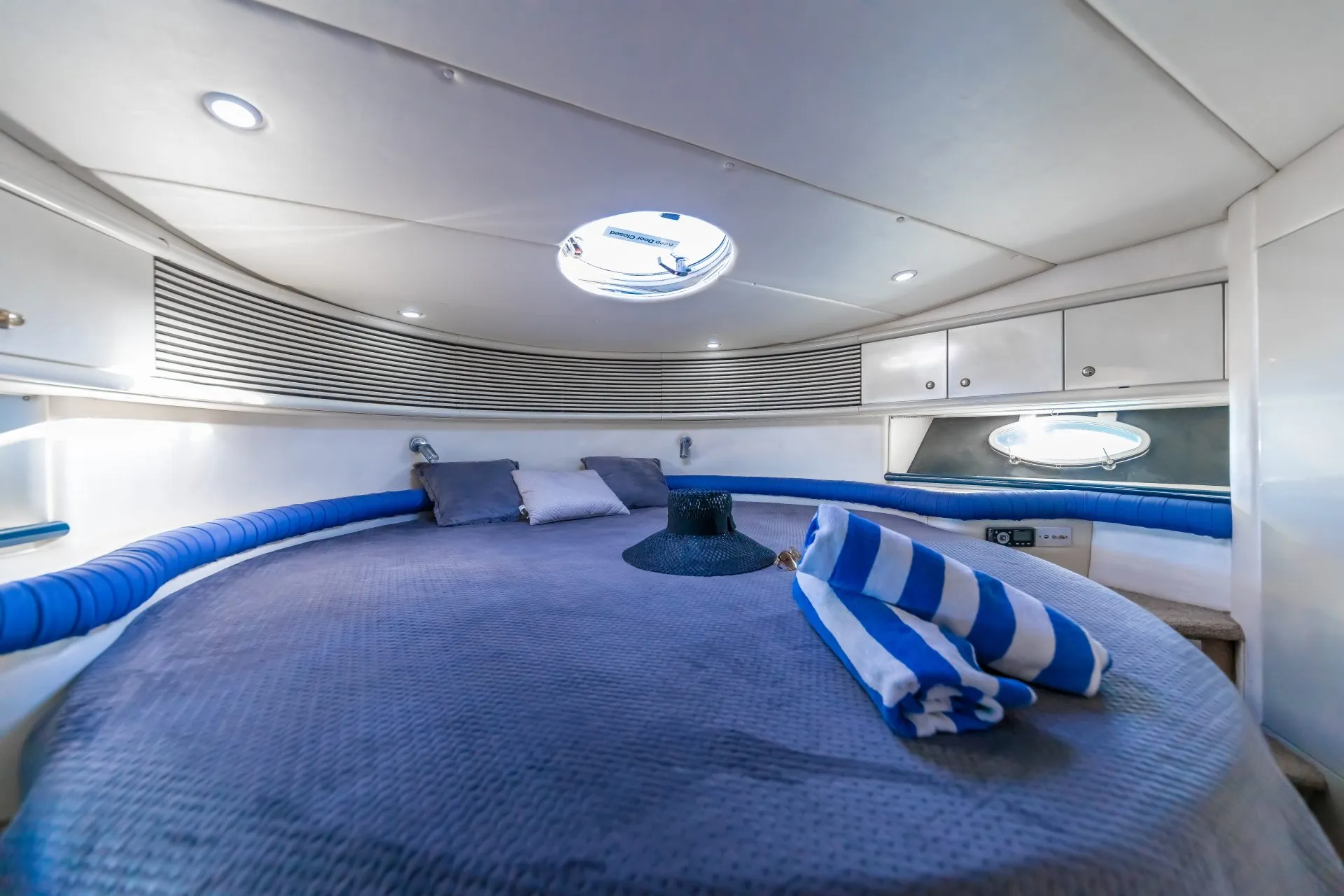 A cozy yacht cabin with a circular bed adorned with blue and grey bedding, pillows, and a striped towel. The cabin features white walls, cabinetry, and a small porthole window letting in natural light. A black hat sits on the bed, and soft ceiling lights illuminate the space. Perfect for those who enjoy Luxury Yacht Rentals Sydney.