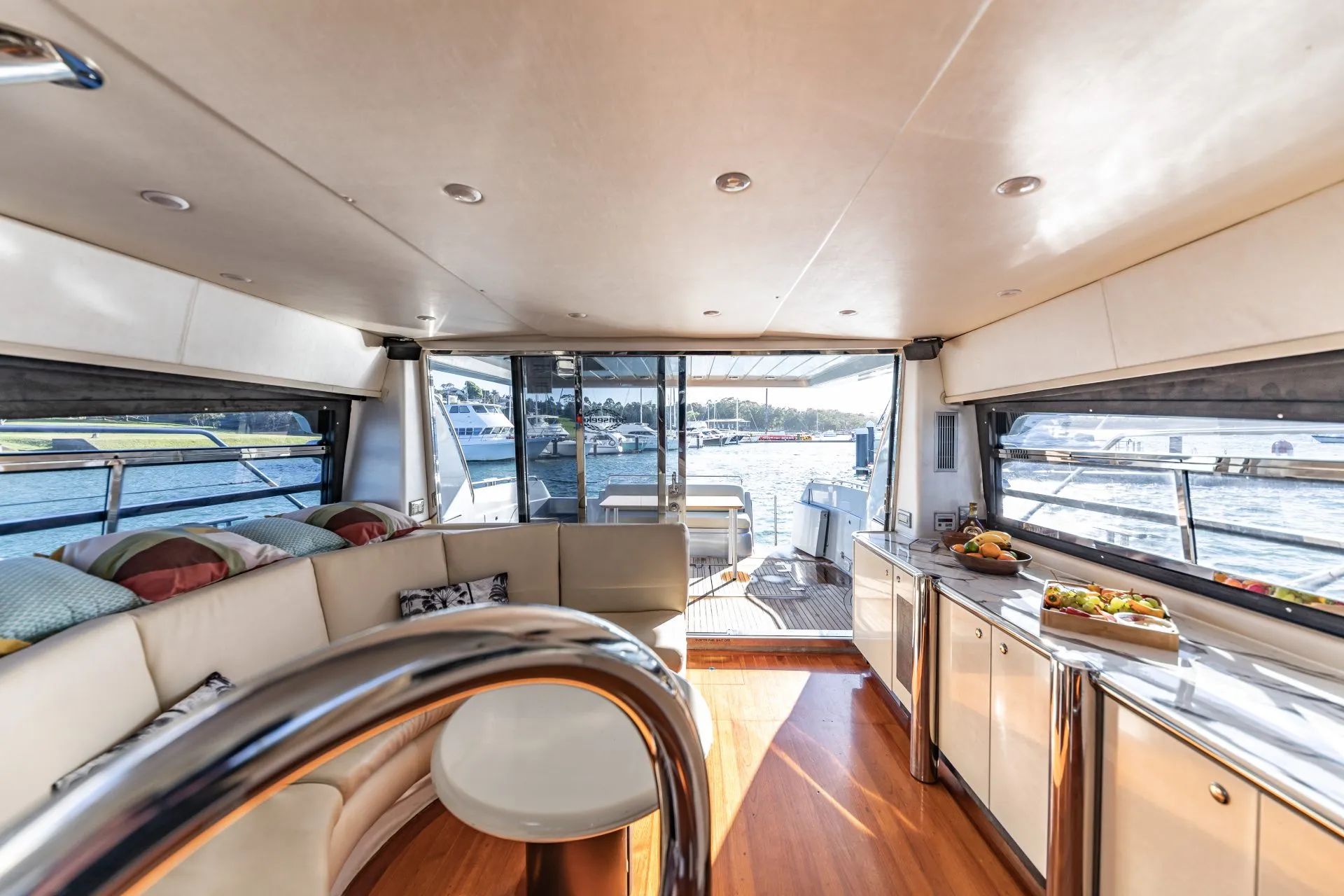 Interior of a luxury yacht featuring a spacious living area with white leather seating, a rounded table, large windows showcasing a marina view, and a small kitchen area equipped with cabinets and a counter displaying plates of food—perfect for The Yacht Social Club Event Boat Charters.