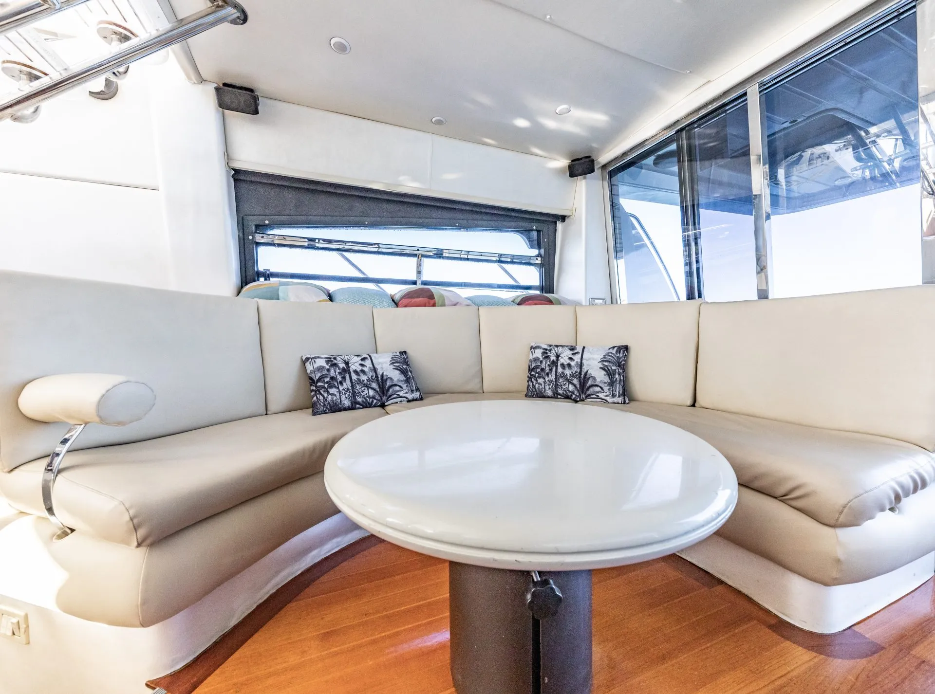 Interior of a yacht featuring a curved, beige leather sofa with tropical-themed cushions surrounding a circular white table. The area is well-lit with natural light from large windows and artificial lighting from the yacht's interior, perfect for events hosted by The Yacht Social Club Event Boat Charters.