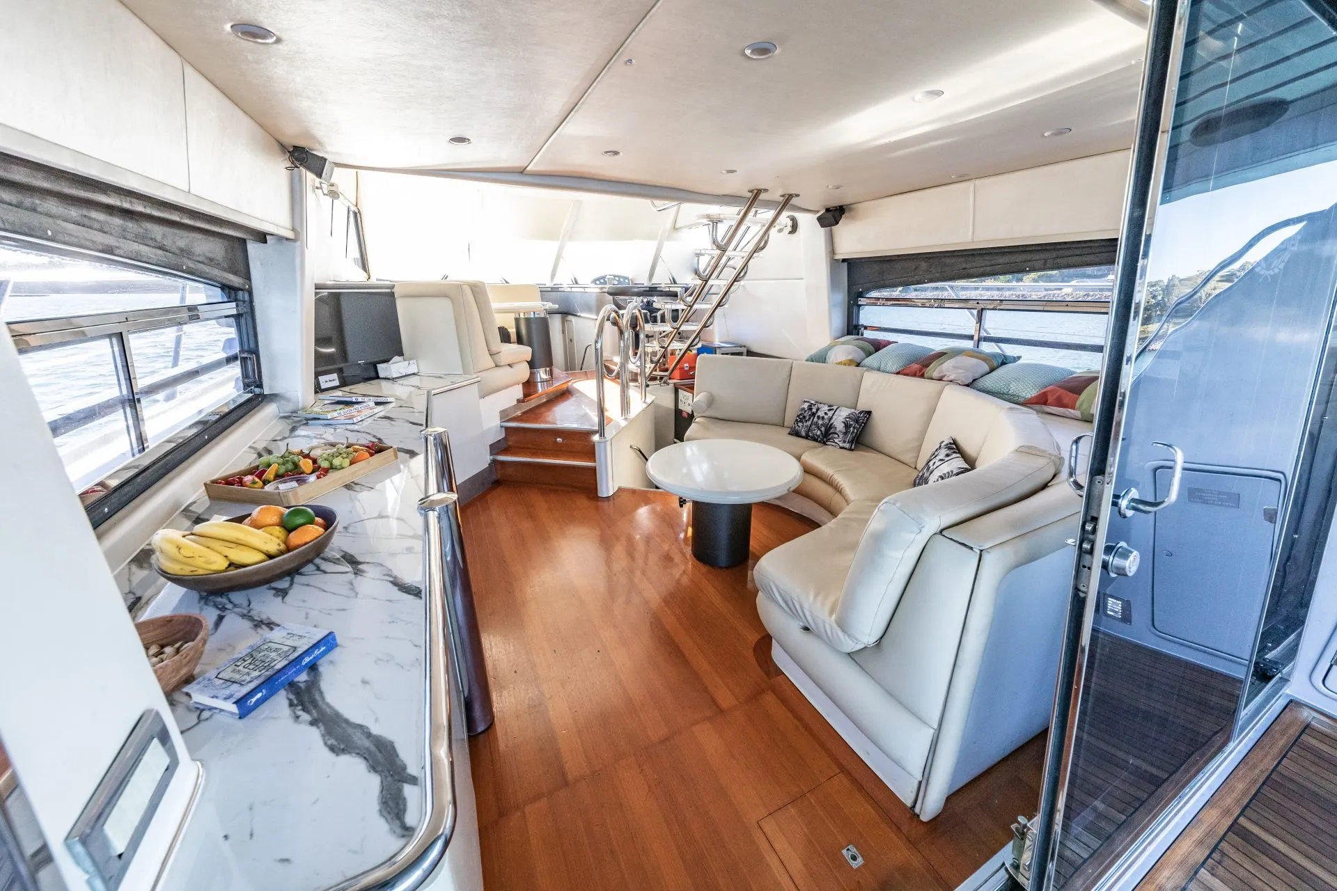 Interior of a luxurious yacht featuring a spacious living area with a white L-shaped sofa, wooden floors, and large windows. The foreground shows a marble countertop with a bowl of fruit and snacks. Stairs lead to an upper deck, illuminated by ample natural light—perfect for Sydney Harbour Boat Hire by The Yacht Social Club.