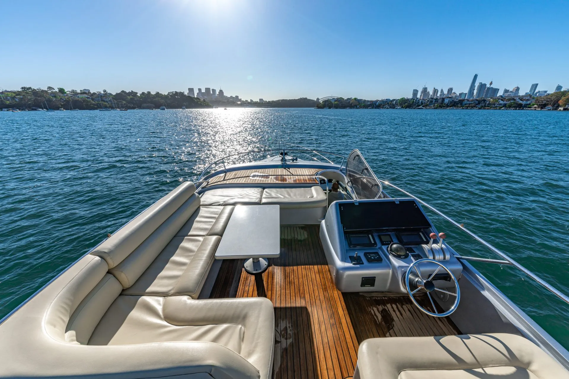 The sun reflects off the calm waters as seen from the deck of a luxurious boat. The Yacht Social Club offers cushioned seating, a table, and navigational equipment. In the distance, Sydney's skyline is visible under a clear blue sky, making for an exquisite experience on Sydney Harbour.