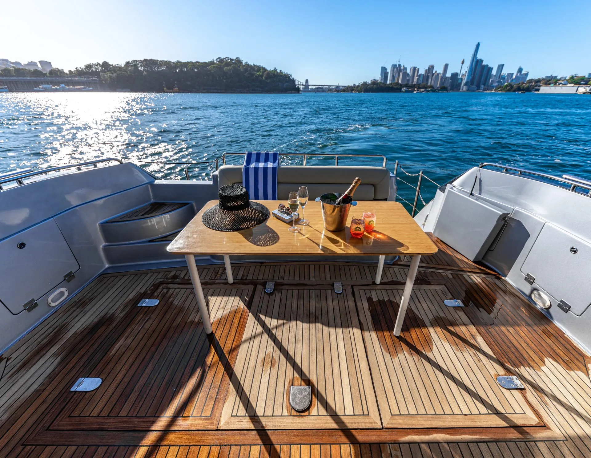 A view from the deck of a luxury yacht rentals sydney shows a table set with drinks, a hat, and a towel against a backdrop of a sunny day on the water. The city skyline and a bridge are visible in the distance. The deck features wooden flooring and gray seating areas.