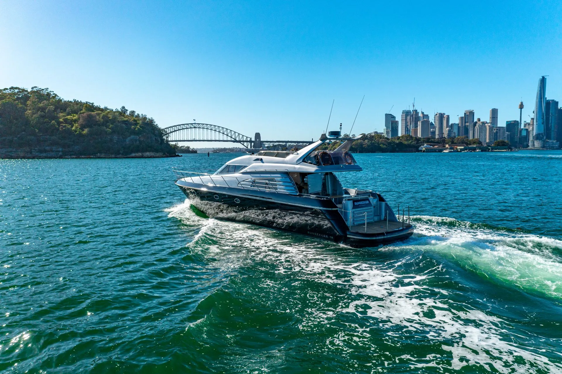 A sleek motor yacht sails through blue-green waters near a coastal city. The backdrop includes a suspension bridge, skyline with tall buildings, and a lush, hilly area to the left. Enjoy Luxury Yacht Rentals Sydney or join The Yacht Social Club for an unforgettable experience. The sky is clear and bright.