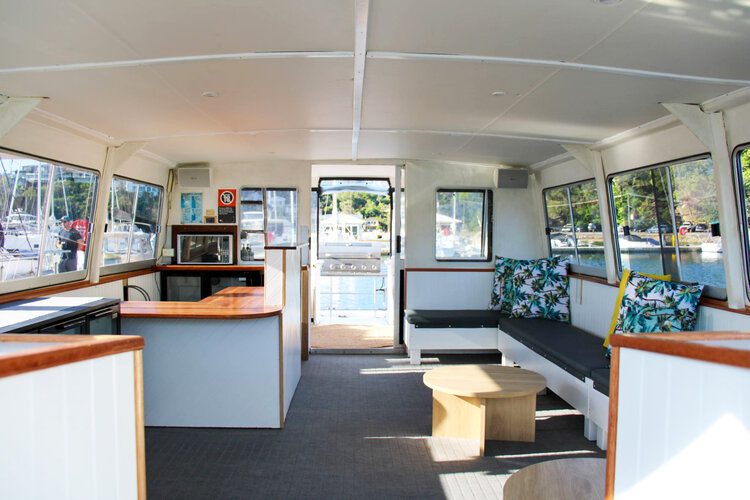 The interior of a boat features a wooden countertop with a microwave, sink, and cozy sitting area. Tropical-themed cushions adorn the seating around a circular wooden table. Large windows provide ample natural light and offer stunning views of the marina outside, perfect for events like The Yacht Social Club in Sydney Harbour.