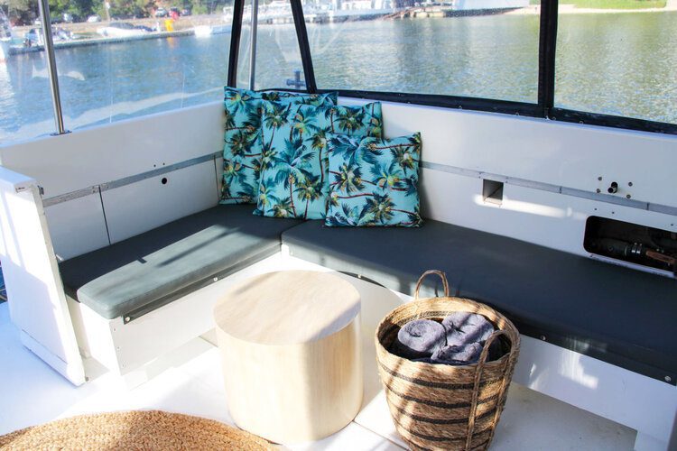 A boat's seating area with blue cushions and tropical-patterned pillows. There's a round, wooden stool in front and a wicker basket with towels to the side. The Yacht Social Club Sydney Boat Hire positions you on a calm body of water with a view of trees and other boats in the background.