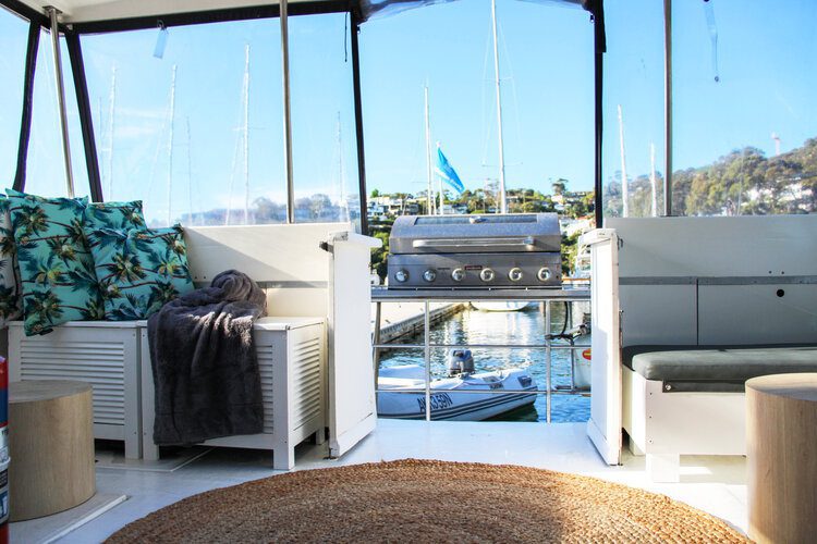 A cozy boat interior featuring a large circular woven rug, cushioned seating with tropical-patterned pillows, and a gray throw blanket. Outside, a dock with sailboats is visible against blue skies and hillside homes. Perfect for Boat Parties Sydney The Yacht Social Club events.