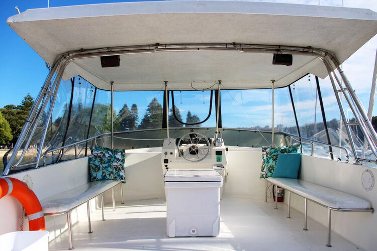 The interior of a boat’s cockpit area is shown, featuring a white steering console, bench seats on each side, a small central white table cooler, and colorful tropical-patterned cushions. Visible are clear enclosures and a life ring. Trees and blue sky are in the background, perfect for Boat Rental and Parties Sydney The Yacht Social Club.