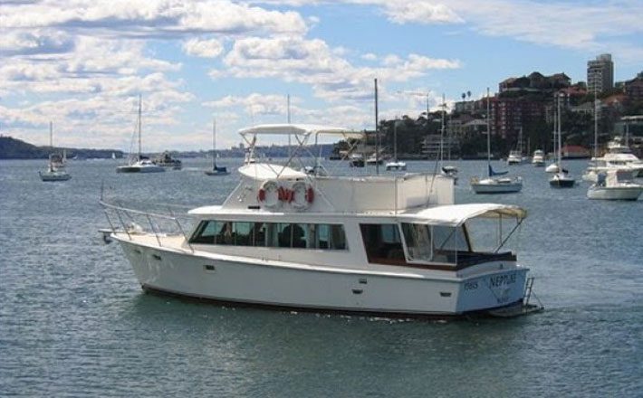 A white motor yacht named "Neptune" is sailing on a calm body of water with several other boats anchored in the background. The sky is partly cloudy, and there are buildings and green hills visible in the distance.