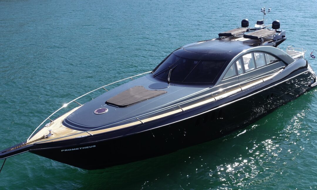 A sleek, modern yacht named "Prometheus" is floating on calm, clear blue water. The yacht features a streamlined design with a black hull and a wooden deck. The sky is clear, suggesting a sunny day.