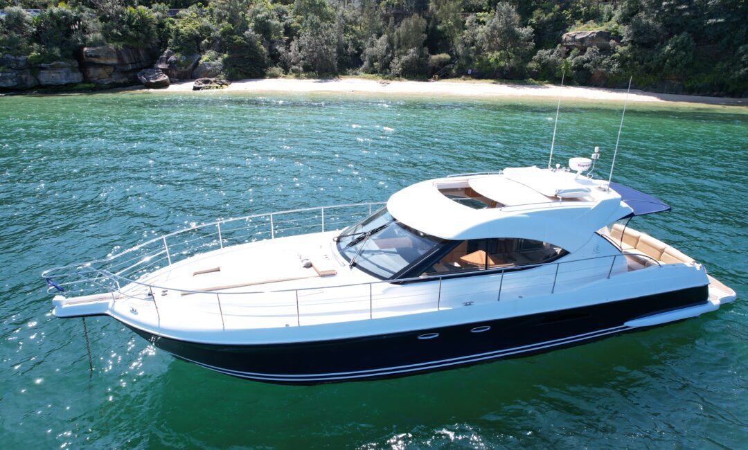 A sleek, white and black yacht floats on clear, green water. The boat has a streamlined design with a covered seating area. In the background, a small sandy beach is surrounded by lush green trees.