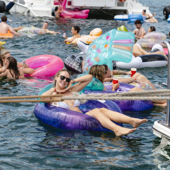 A group of people enjoy a sunny day on the water, lounging on colorful inflatable tubes and floats. They are smiling and relaxed, surrounded by boats in the background.