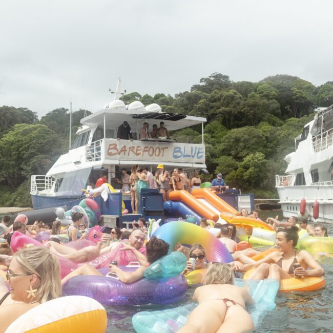 A lively scene of people enjoying a party on colorful inflatable floats in the water. Two boats are nearby, one with a "Barefoot Blue" banner. The area is surrounded by lush greenery under an overcast sky.