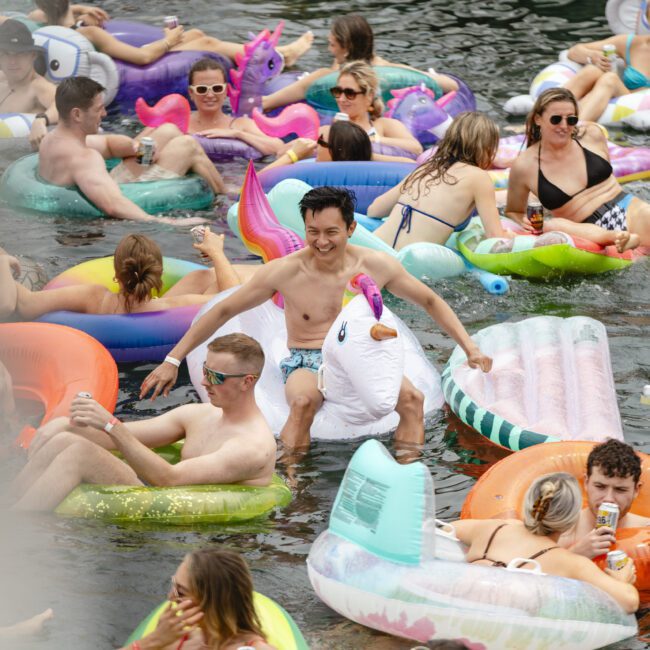 A crowded river scene with people floating on colorful inflatables like swans, unicorns, and pizzas. They are enjoying a sunny day, with many wearing swimwear and holding drinks. The atmosphere is lively and festive.