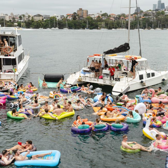 A lively scene of people enjoying a boat party in a harbor. Dozens of individuals are floating on colorful inflatables near two anchored boats, with a city skyline and bridge visible in the background. The atmosphere is fun and festive.