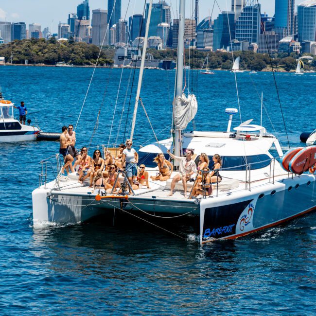A group of people are enjoying a sunny day on a sailing catamaran in the water, with a city skyline visible in the background. Some are sitting and others are standing on the deck.