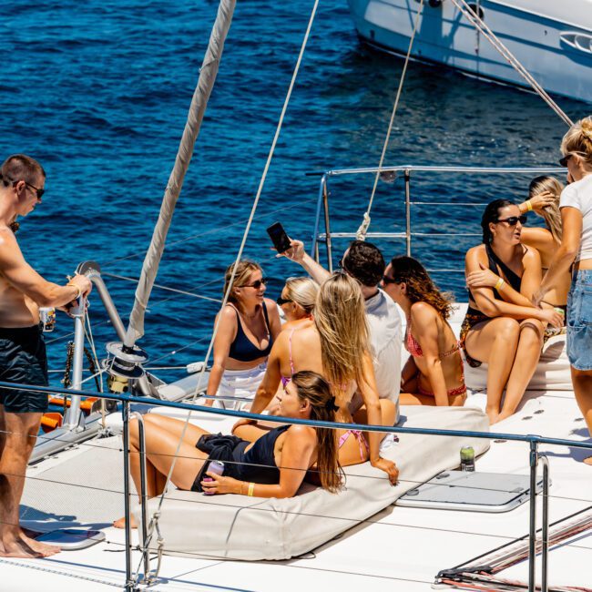 A group of people relaxing on a yacht in sunny weather. Some are sitting, while others are reclining on cushions. The ocean is visible in the background, and another yacht can be seen nearby.
