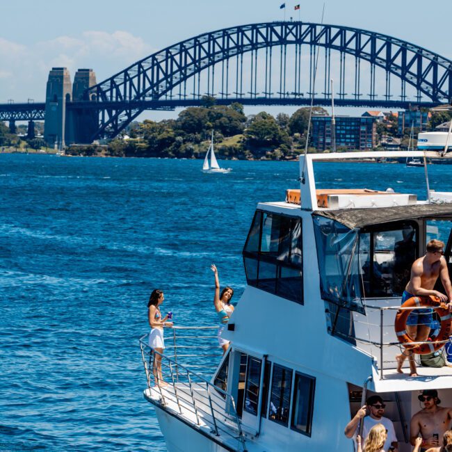 A boat with people enjoying the sunny day floats on Sydney Harbour, with the iconic Sydney Harbour Bridge in the background. A few people are standing on the deck, one waving, and a sailboat is seen in the distance.