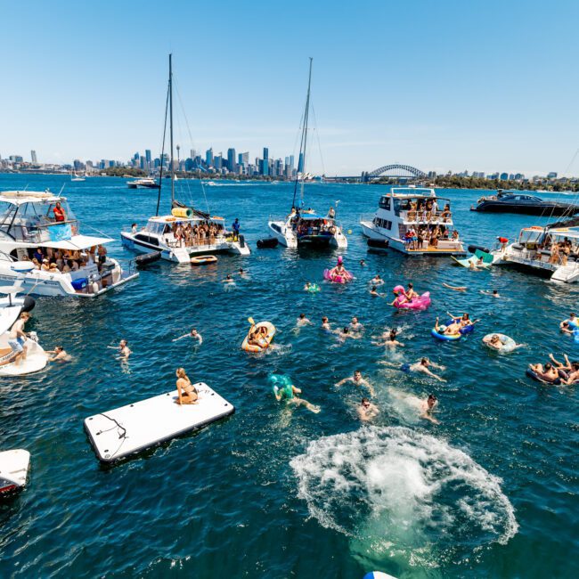 A lively scene on the water with numerous boats and people swimming and lounging on inflatables. The city skyline and a bridge are visible in the background. The water is bright blue, capturing a sunny day.