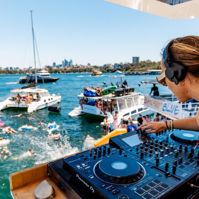 A woman DJ is mixing music on a boat overlooking a lake party. People are swimming and enjoying the water, with other boats and a city skyline visible in the background. It's a sunny day, creating a festive atmosphere.