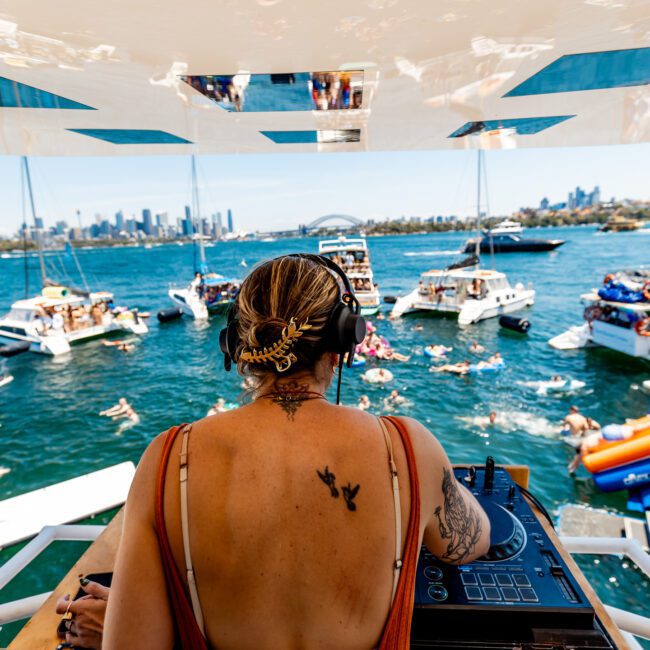 A DJ with tattoos is playing music on a boat during a lively party in a bay. The scene includes several boats nearby and people swimming and relaxing in the water under a sunny sky. The city skyline is visible in the background.