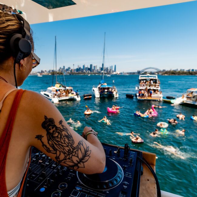 A DJ with headphones plays music on a yacht overlooking a lively pool party. People are floating on inflatables in the water. The city skyline with tall buildings is visible in the background under a clear blue sky.