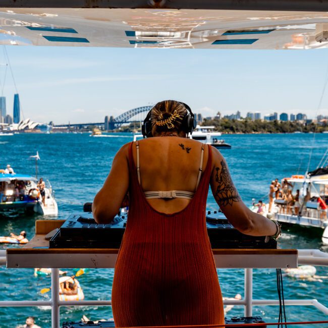 A DJ wearing headphones performs on a boat with a scenic backdrop of a city skyline and a bridge. Several boats and swimmers are visible on the blue water under a clear sky.