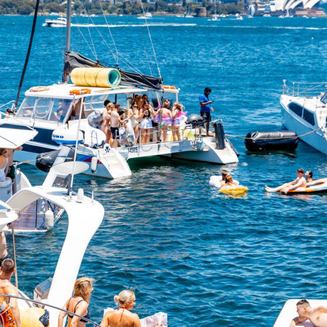 A group of people enjoying a sunny day on boats in a harbor, with music and pool floats. The city skyline is visible in the background. Several individuals are swimming or lounging on inflatable chairs in the water.