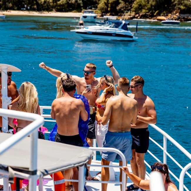 A group of people in swimsuits are enjoying themselves on the deck of a yacht, dancing and socializing. The yacht is surrounded by clear blue water, with trees and a distant boat in the background.