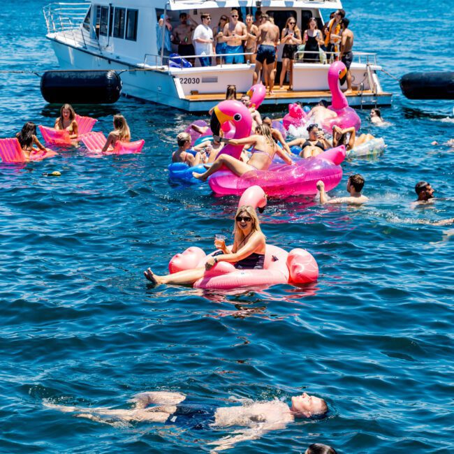 A lively scene on a sunny day with people enjoying a yacht party. Many are floating on colorful inflatable toys, including flamingos, in the water near the boat. The sky is clear, and the backdrop includes a distant city skyline.
