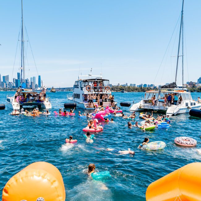People enjoying a sunny day on the water with floats. Several boats are anchored nearby, with the city skyline in the background. The scene is lively, with colorful inflatable tubes and swimmers in bright blue water.