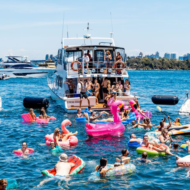 People enjoying a sunny day on the water, with a large boat in the background. Many are floating on colorful inflatables, including pink flamingos and various pool floats, surrounded by others swimming and relaxing in the blue water.