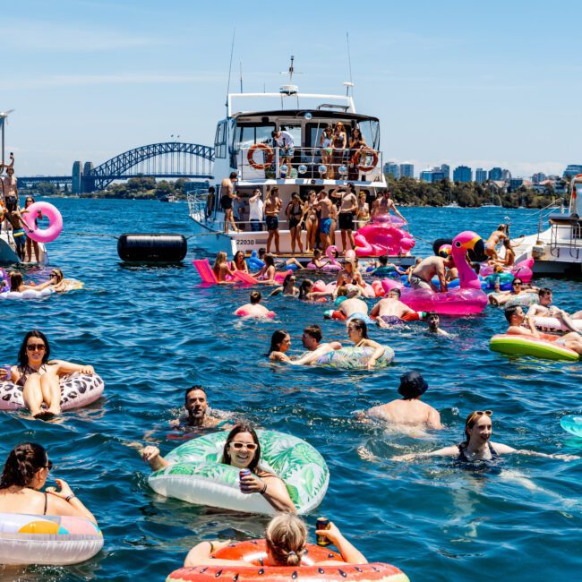 People enjoying a sunny day on the water with inflatable rafts and floats. A yacht is in the background with more people on board. The Sydney Harbour Bridge and city skyline are visible in the distance.
