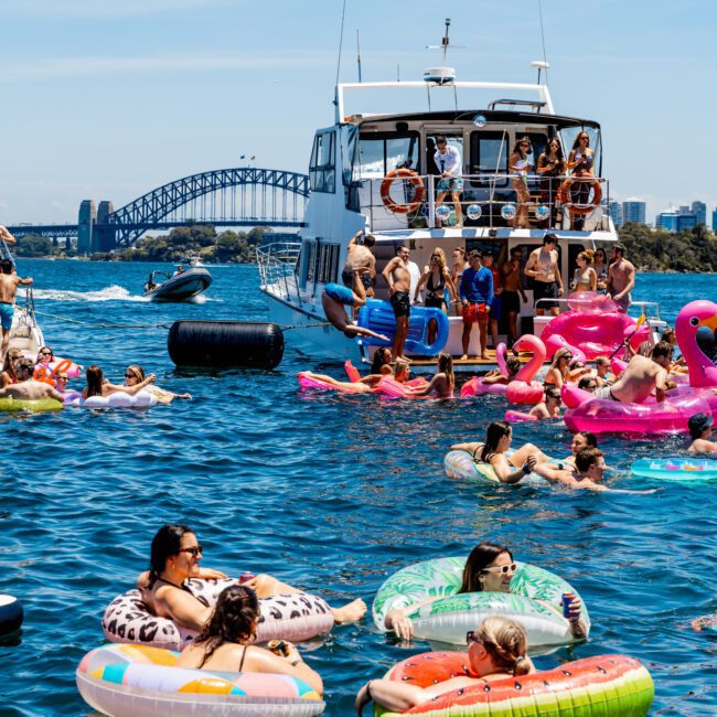 People enjoying a sunny day on a large body of water, lounging on colorful inflatable tubes and floats near a yacht. A bridge and city skyline are visible in the background, under a clear blue sky.