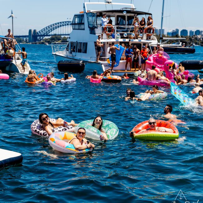 People enjoy a sunny day on the water, floating on colorful inflatable loungers near a large boat. The scene is lively with a mix of boats and socializing in the background.