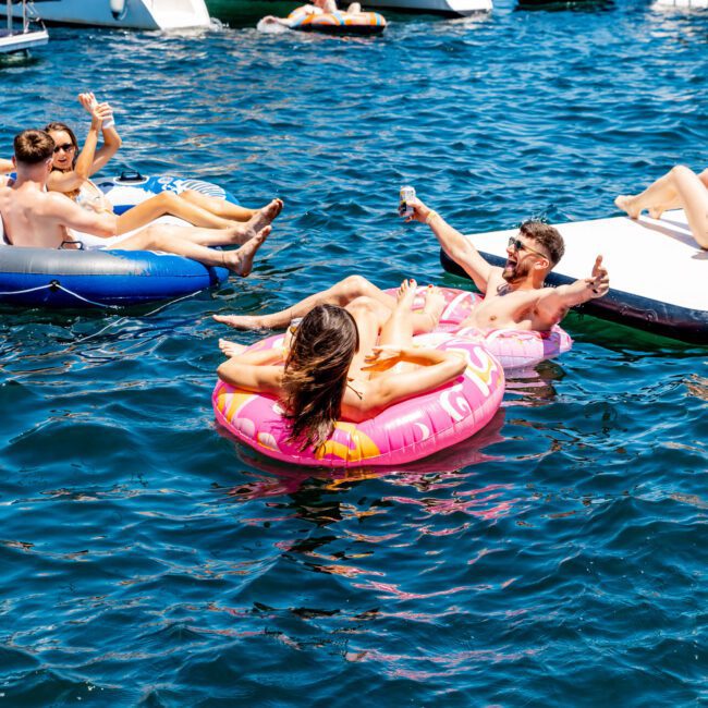 A group of people relax on inflatable tubes in a sunny, bustling harbor with boats in the background. They are casually enjoying drinks and the water, with a city skyline visible in the distance.