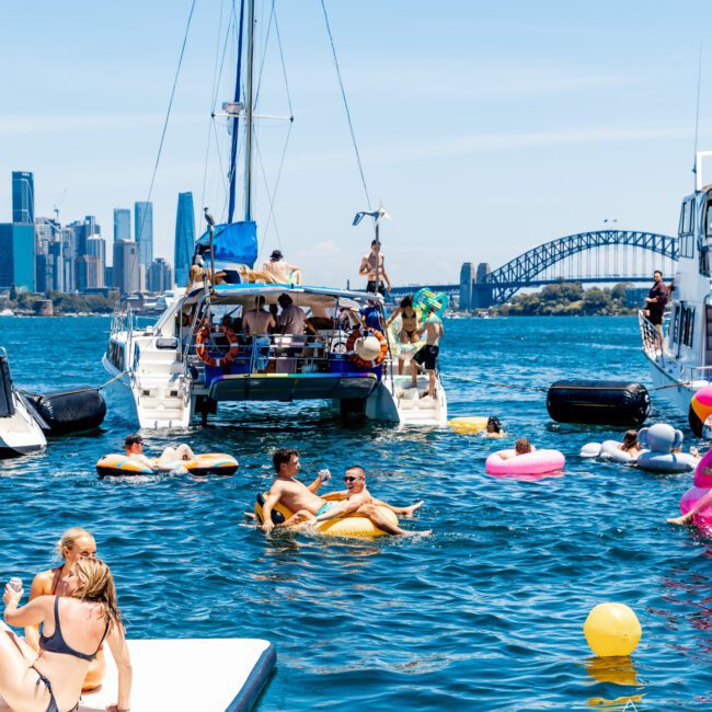 People are enjoying a sunny day on the waterfront, with several boats anchored nearby. Some are swimming or floating on inflatables. A city skyline and a bridge are visible in the background under a clear blue sky.