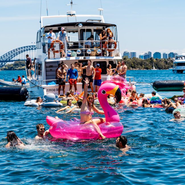 A lively scene on the water with people enjoying a yacht party. Some are swimming, while others relax on inflatable floats, including a pink flamingo. A yacht and city skyline are visible in the background under a clear blue sky.