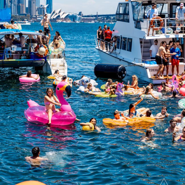 A lively scene with people swimming and floating on inflatables near boats in a bay. A woman sits on a pink flamingo float. The city skyline and Opera House are visible in the background. It's a sunny day with a festive atmosphere.