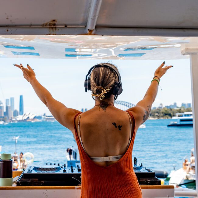 A DJ wearing headphones performs on a boat with arms raised, facing the open water. The city skyline and boats are visible in the background, under a sunny sky. Speaker systems flank the DJ. A small butterfly tattoo is visible on the DJ's back.